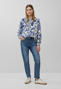 Chemisier à manches longues à fleurs bleu et blanc, associé à un jean bleu clair et des baskets blanches avec des semelles brunes. Le chemisier possède un col bouton.