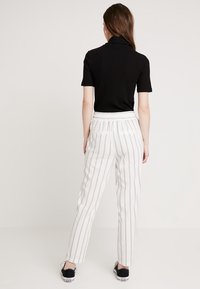 Kvinna som står med ryggen mot kameran iförd en svart kortärmad topp, vita byxor med vertikala ränder och svarta sneakers mot en neutral bakgrund.