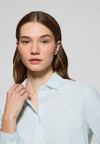 Camicia button-up a righe blu chiaro con una texture morbida, colletto e logo sottile sul petto. Presenta bottoni bianchi classici.