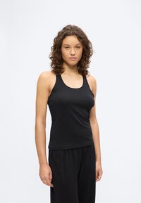 Νέα γυναίκα με κατσαρά μαλλιά, που φοράει μαύρο αμάνικο τοπ και φαρδιά μαύρη παντελόνια, στέκεται μπροστά σε απλό λευκό φόντο.