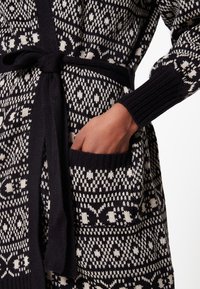 Černo-bílý pletený kardigan s geometrickým vzorem, zavazovacím páskem a kapsou na pravé straně. Texturovaná tkanina a žebrované manžety.