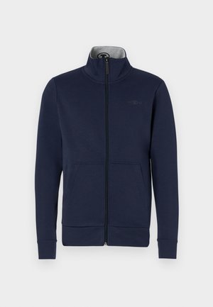Marineblauwe ritsjack van zacht materiaal, met een staande kraag, twee voorzakken en geribde boorden en zoom. Subtiele logo-details op de borst.