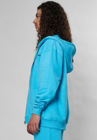 Svetlomodrá mikina na zips vyrobená z mäkkej látky, s kapucňou, dlhými rukávmi a rebrovanými manžetami a lemom. Minimalistický dizajn s detailom loga.