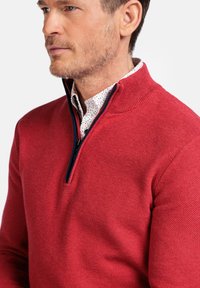Rode gebreide pullover met een hoge kraag en een rits tot halverwege. Heeft een gestructureerd patroon en marineblauwe accenten. Draagt over een gebloemd shirt.
