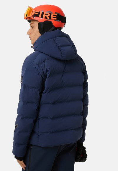 Navy blue puffer jacket with a hood, featuring quilted sections, a logo on the sleeve, and a smooth texture. Worn with a bright orange ski helmet.