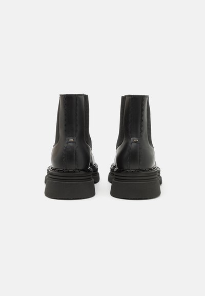 Bottes noires à chevilles avec une finition en cuir lisse, des panneaux latéraux élastiques et une épaisse semelle en caoutchouc texturée. Comprend des coutures décoratives à l'arrière.