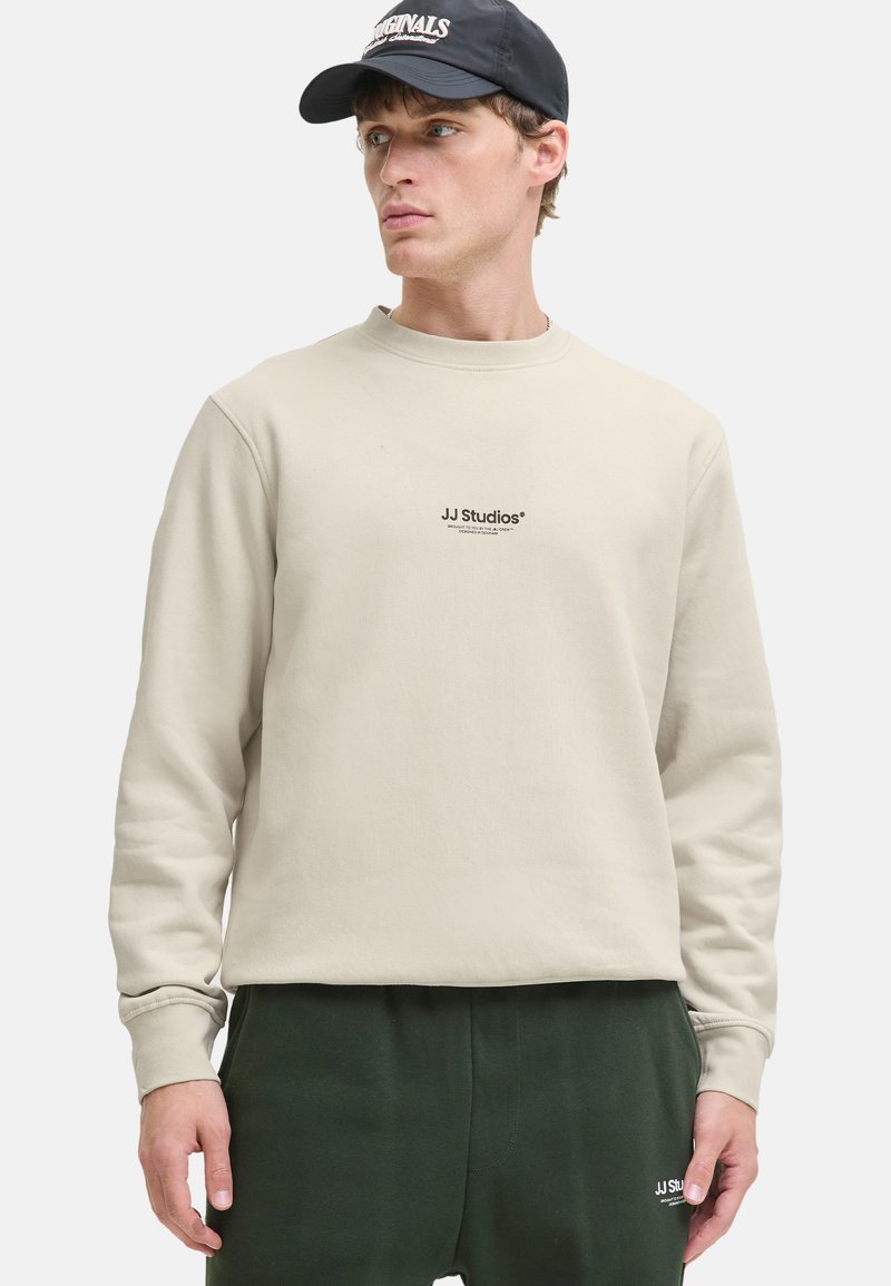 Sweat-shirt beige avec col rond et manches longues, arborant un logo noir "JJ Studios" sur la poitrine. Porté avec un pantalon de jogging vert foncé.