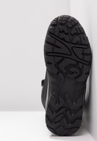 Semelle noire de botte de randonnée avec des crampons profonds et texturés, inclinée contre un fond blanc en coin.