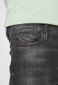 Vaqueros de mezclilla negra con un acabado texturizado y un ligero desgaste. Presenta una etiqueta de marca visible en el bolsillo trasero y un diseño estándar de cinco bolsillos.
