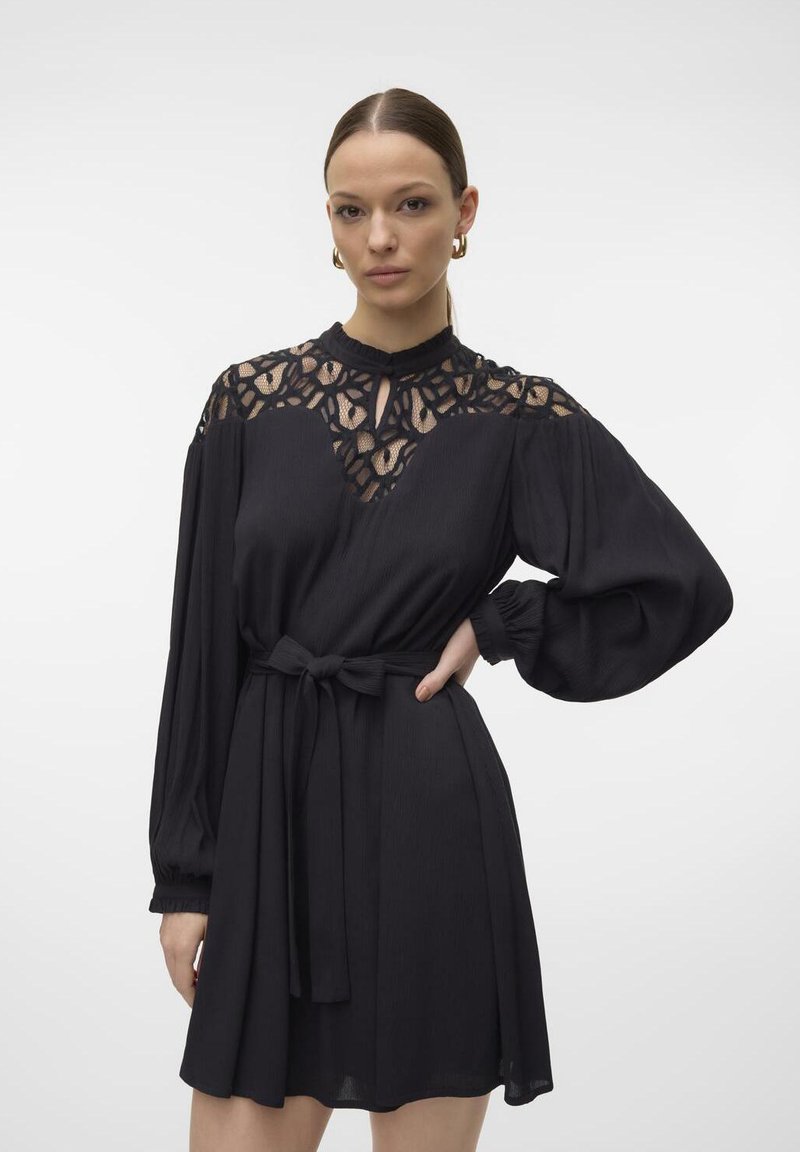 Rochie neagră cu mâneci lungi, panou din dantelă la guler, textură plisată și o curea din material, având un guler înalt și o fustă evazată.