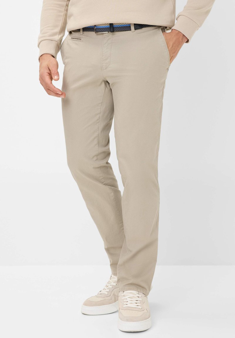 Beige Baumwoll-Chinos mit geradem Schnitt, subtiler Textur und seitlichen Taschen. Kombiniert mit hellen Sneakers und einem kontrastierenden blauen Gürtel.