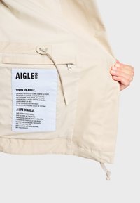Poche intérieure d'une veste beige avec une grande étiquette blanche affichant la marque AIGLE 1853 et un poème bilingue sur la nature et la liberté, tenue ouverte par une main.