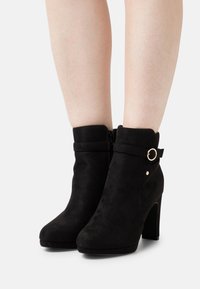 Schwarze Wildleder-Ankle-Boots mit hohen Absätzen, seitlichem Reißverschluss, Riemen mit goldfarbener Ringbeschläge und glatter Textur.