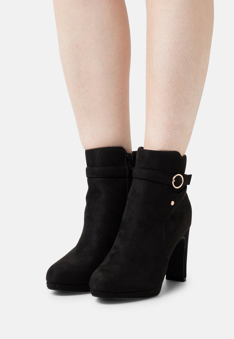 Schwarze Wildleder-Ankle-Boots mit hohen Absätzen, seitlichem Reißverschluss, Riemen mit goldfarbener Ringbeschläge und glatter Textur.