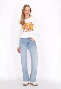 Felpa bianca a maniche lunghe con grafica "FALL VIBES" in arancione e marrone, abbinata a jeans dritti azzurri e sneakers bianche.