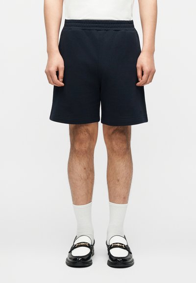 Homme portant un short noir, des chaussettes blanches montantes et des mocassins noirs et blancs avec un détail chaîne, debout devant un fond uni.