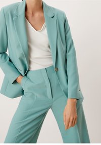 Costume vert bleuâtre clair composé d'un blazer taillé avec un seul bouton et d'un pantalon assorti, porté sur un haut blanc à col en V.