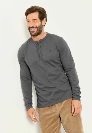 Lächelnder Mann, der an eine Wand lehnt und ein dunkelgraues Langarm-Henleyshirt sowie hellbraune Cordhosen trägt.