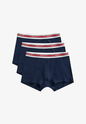 Trois boxers pour hommes bleu marine avec des ceintures blanches ornées d'une bande rouge et du nom de la marque "GANT" répété en blanc.
