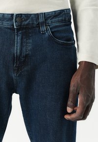Vaqueros de lavado oscuro hechos de denim, con un diseño clásico de cinco bolsillos, cierre de botón y un desgaste sutil en los muslos.