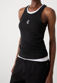 Svart ärmlös ribbad topp över en vit linne, bärs med svarta jeans, visar överkropp och armar mot en enkel bakgrund.