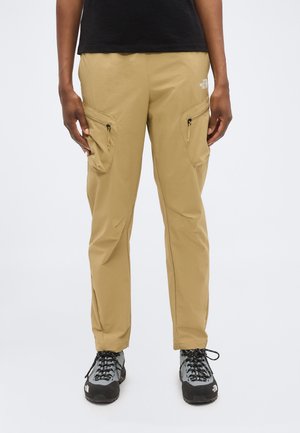 Άτομο που φοράει μπεζ παντελόνι cargo με φερμουάρ στις πλαϊνές τσέπες και μαύρα ορειβατικά παπούτσια, στέκεται μπροστά σε απλό λευκό φόντο.