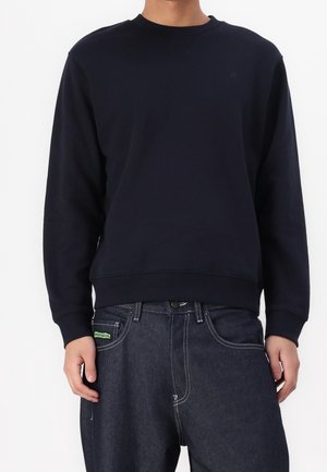 Personne portant un sweat-shirt noir uni à manches longues et un jean bleu foncé, debout avec les bras détendus le long du corps.