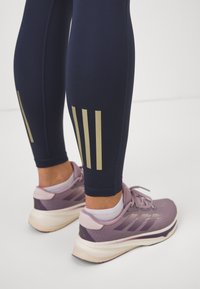 Marinblå leggings med guldtre-randsdesign vid vaden, i kombination med lila träningsskor som har lätta detaljer och en strukturerad meshovandel.