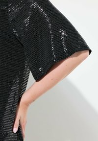 Top negro con un diseño holgado y de manga corta, con una superficie texturizada y de lentejuelas que refleja la luz, realzando el interés visual.
