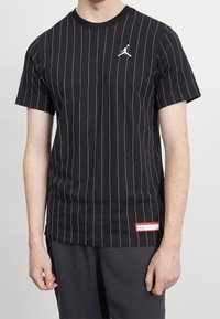 Muž v černém tričku s krátkým rukávem a bílými vertikálními pruhy, s bílým logem na hrudi a červeným obdélníkovým štítkem blízko lemu.