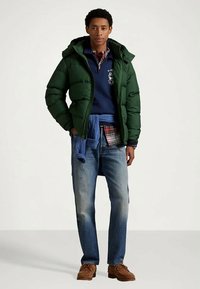 Νεαρός άνδρας φορώντας πράσινο μπουφάν, ναυτικό πουλόβερ, καρό πουκάμισο, μπλε τζιν και καφέ παπούτσια, που στέκεται μπροστά σε απλό φόντο.