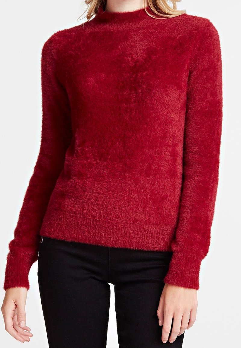 Femme portant un pull rouge duveteux à manches longues et un pantalon noir, debout les bras détendus le long du corps.