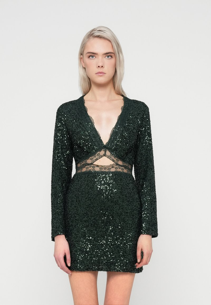 Σκούρο πράσινο, μίνι φόρεμα με παγιέτες και μακριά μανίκια, με βαθύ V-ντεκολτέ και δαντέλες στη μέση, παρουσιάζοντας μια υφή με λάμψη.