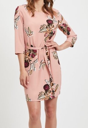 Femme portant une robe fleurie rose jusqu'aux genoux, manches trois-quarts et ceinture en tissu nouée à la taille.