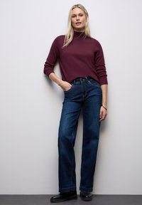 Maglione a collo alto color bordeaux abbinato a jeans a gamba larga di un blu scuro. L'ensemble presenta un design semplice con una trama liscia e dettagli minimi.