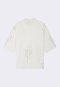 Camisa blanca de manga corta con patrones de cactus negros, tela texturizada y cuello. La vista trasera muestra un detalle de la costura central.