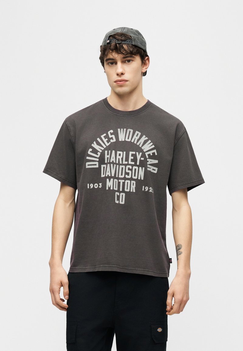 Giovane uomo che indossa una maglietta Dickies Workwear Harley-Davidson nera scolorita e un cappello grigio indossato al contrario, in piedi davanti a uno sfondo semplice.