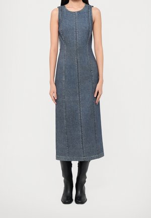 Robe midi en jean ajustée sans manches avec des coutures verticales, portée avec des bottes noires mi-mollet sur une personne debout devant un fond uni.