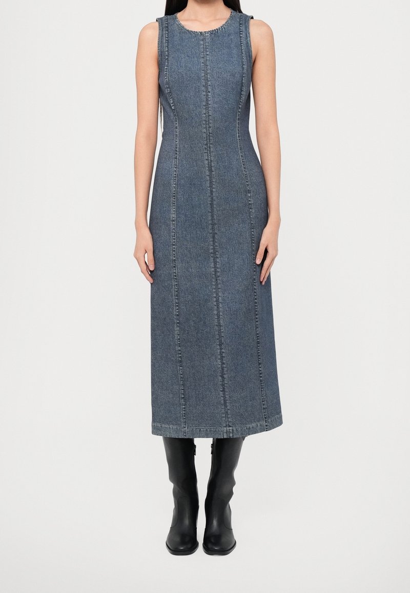 Robe midi en jean ajustée sans manches avec des coutures verticales, portée avec des bottes noires mi-mollet sur une personne debout devant un fond uni.