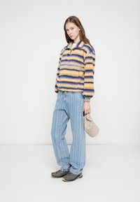 Pulêover felpudo multicolorido com riscas amarelas, roxas e azuis; combinado com jeans azul claro às riscas e ténis cinzentos. Segura uma mala bege.