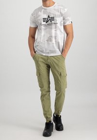 Biała koszulka w kamuflażu z czarnym logo, w zestawie z oliwkowymi spodniami cargo z kieszeniami oraz czarnymi trampkami.
