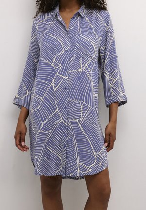 Vrouw draagt een loszittende blauw shirtjurk met een wit abstract bladmotief en lange mouwen tegen een effen lichte achterwand.