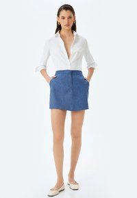 Chemise blanche à boutons, mini-jupe texturée bleu avec poches, et ballerines beiges, présentant une combinaison de tenue soignée et décontractée.
