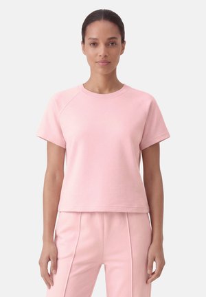 Donna in piedi di fronte, indossa una maglietta rosa chiaro a maniche corte e pantaloni abbinati su uno sfondo chiaro uniforme.