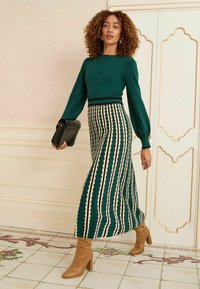 Maglione verde lavorato a maglia con maniche a sbuffo, abbinato a una gonna midi a righe con motivi a scallop color crema. Stivali marroni con tacco e borsa nera.