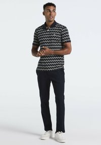 Polo à motifs noir et blanc avec des manches courtes, associé à un pantalon bleu marine et des baskets blanches. Le design présente des formes de losanges.