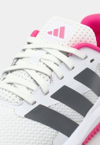 Chaussure de sport blanche avec des accents roses, dotée d'un dessus perforé, de rayures grises et de lacets blancs. Semelle texturée avec des détails roses.