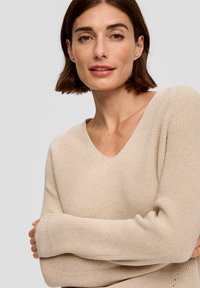 Beżowy sweter prążkowany z dekoltem w kształcie litery V i wydłużonymi rękawami. Tekstura dzianiny jest luźna i miękka, z delikatnymi akcentami na końcu. 