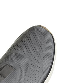 Primer plano de la puntera de un zapato deportivo de tela gris tejida con agujeros de ventilación y un borde de goma beige sobre una suela blanca.