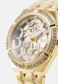 Montre analogique en or avec un cadran transparent, des engrenages complexes et trois sous-cadrans. Présente une lunette ornée de cristaux et un design de bracelet.